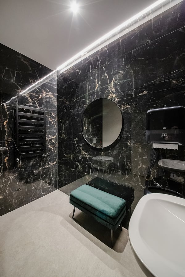 Luxurious bathroom with black marble walls, round mirror, modern fixtures, and a green velvet bench.