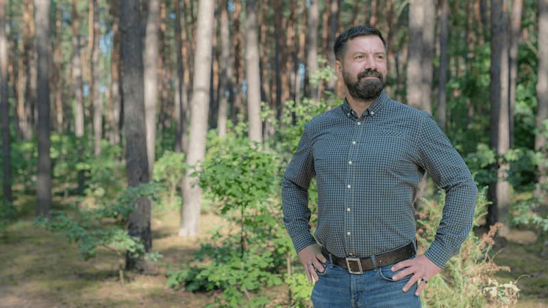 Maciej stands in a sunlit forest wearing a checkered shirt and jeans.