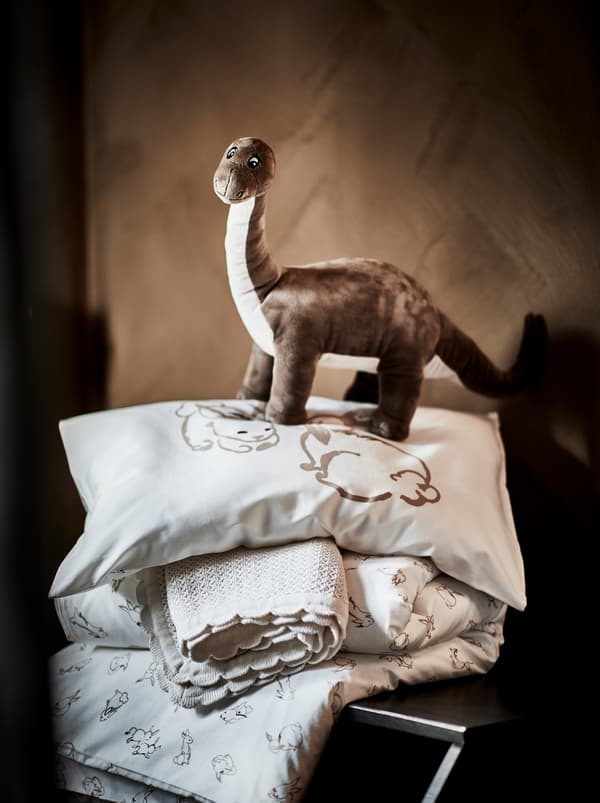 Mainan lembut dinosaur JÄTTELIK berwarna coklat berdiri di atas bantal dan selimut dengan linen katil dalam ton warna semula jadi yang lembut.