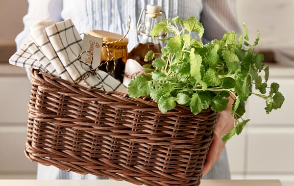 Mâini care oferă un coș GABBIG plin cu șervete, conserve făcute în casă în borcane de sticlă și ierburi proaspete.