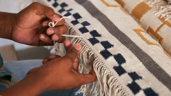 Mains tissant la frange d’un tapis dans des tons de blanc, bleu foncé, gris et moutarde. 