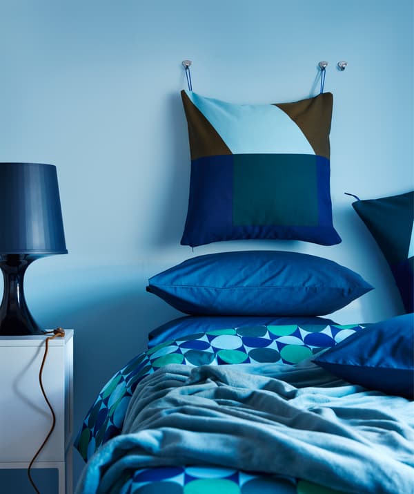 หมอนอิงที่มีสีฟ้าและสีเขียวจากปลอกหมอนอิง MAJALISA ของอิเกียแขวนอยู่ที่ผนังห้องนอนในห้องนอนที่มีผ้าทอสีฟ้า