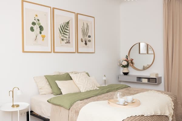 Mājīga guļamistaba ar divguļamu gultu, gaišu tekstilu, dekoratīviem spilveniem, mākslas darbiem pie sienas un apaļu spoguli virs galdiņa.