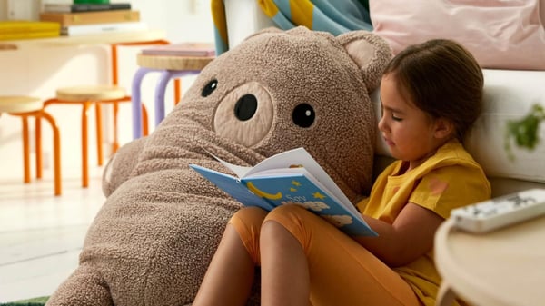 Malá holčička, která sedí vedle velkého plyšového medvěda a čte si knihu. Holčička má na sobě žluté tričko a oranžové kalhoty. Kniha, kterou drží, má modrý obal s obrázky hvězd a mraků. Scéna se odehrává v dětském pokoji nebo obývacím pokoji, kde jsou vidět i další dětské stoličky a nábytek.