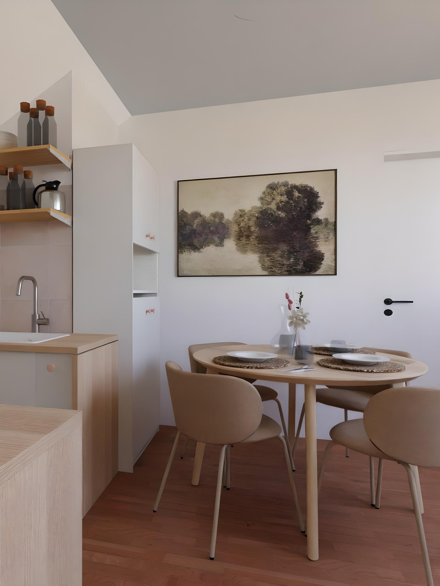 Mala kuhinja i blagovaonica s okruglim stolom za četiri osobe, postavljenim tanjurima i čašama; na zidu slika pejzaža, drveni pod, bijeli zidovi, sudoper, polica s bocama i ormarići.