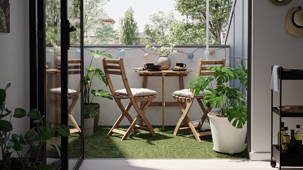 Mali balkon s drvenim stolom i stolicama, podne obloge s veštačkom travom, kao i biljka monstera u beloj saksiji.