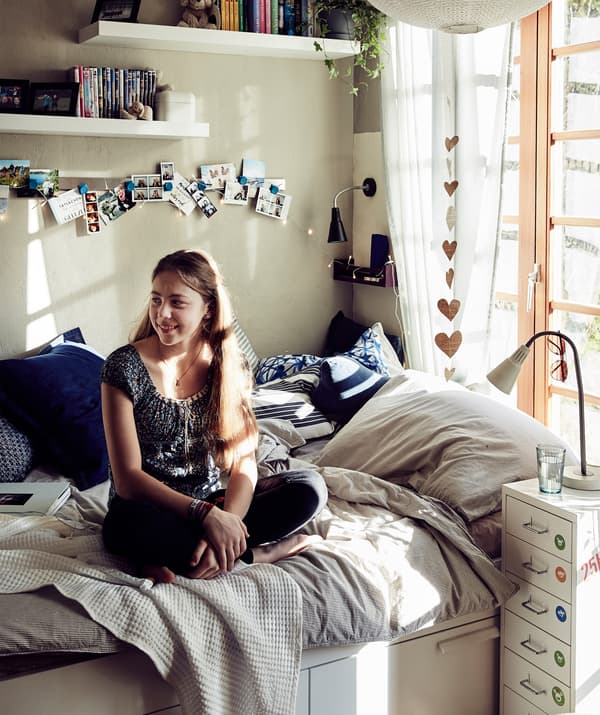 Malin นั่งอยู่บนเตียงที่มีเครื่องนอนสีพื้น หมอนอิงสีน้ำเงินและชั้นวางอยู่บนผนัง