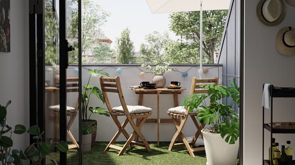 Malý balkon s dřevěnými stoly a židlemi, na zemi je umělá tráva a rostlina Monstera v bílém květináči.