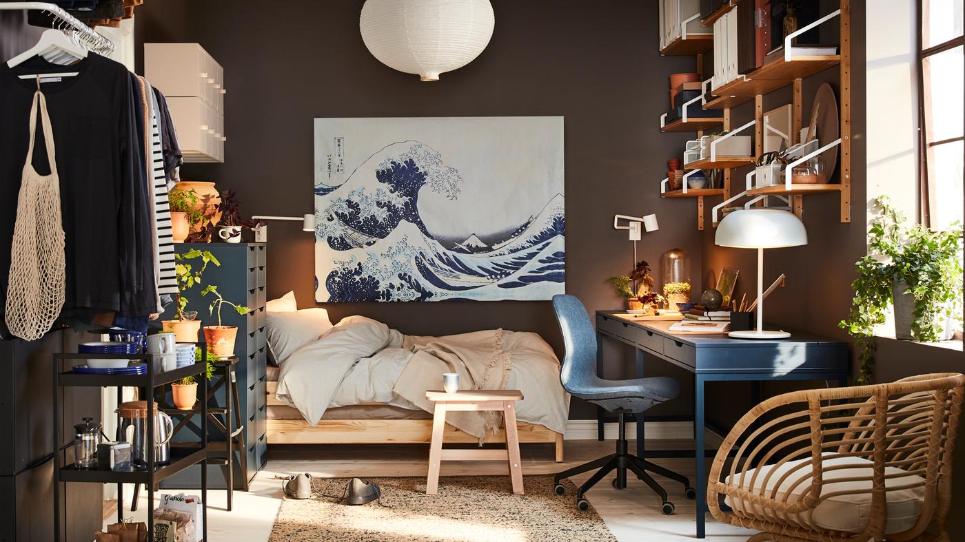 Malý byt s tmavě modrým stolem, ratanovým křeslem, bílým stínidlem závěsné lampy a obrazem s modrou vlnou.