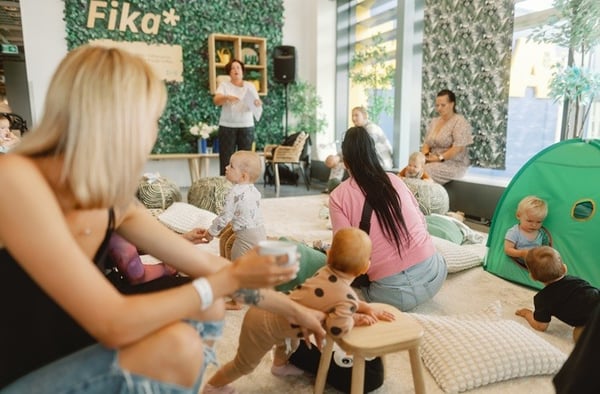Мамы с малышами смотрят на ведущего в специально оборудованной зоне шведского ресторана во время родительских утренников. Комната оформлена в светлом дереве и песочных тонах.