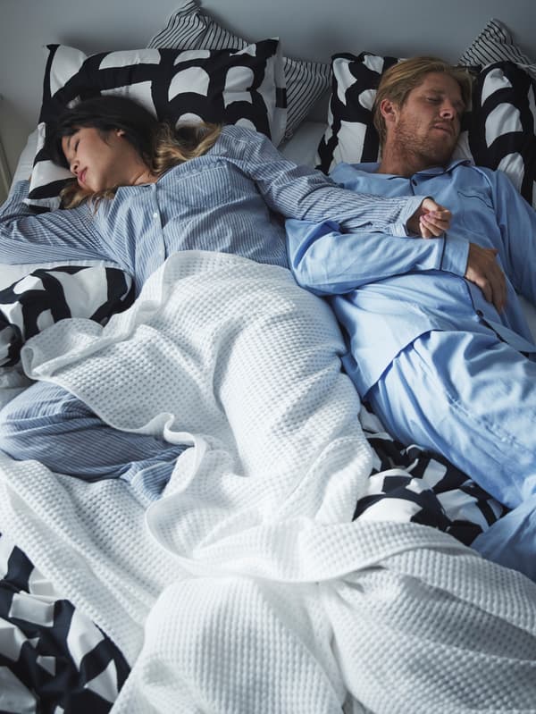 Man and woman asleep in bed wearing pyjamas with ruffled sheets, duvet, pillows and VÅRELD bedspread.