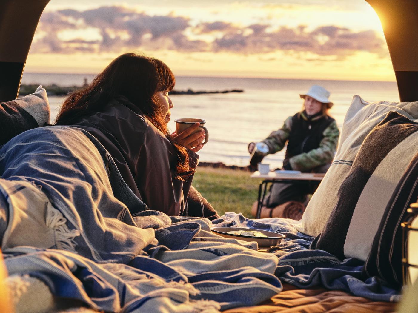 Man blickt aus einem Autokofferraum auf einen See. Eine Frau liegt im Kofferraum, der zum Travel-Van ausgebaut wurde, trinkt eine Tasse Heißgetränk. Ein Mann sitzt vor dem See an einem Klapptisch, mit Fischer-Hut und ebenfalls Heigetränk.