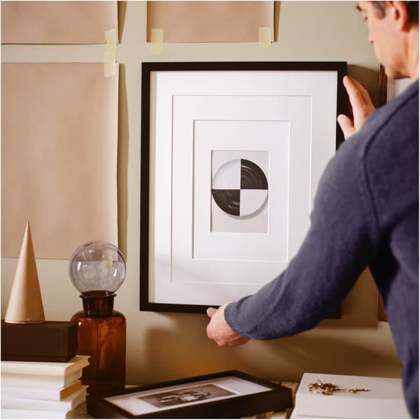 Man hangs a picture in a black RIBBA frame on a beige wall onto which brown paper templates have been taped.