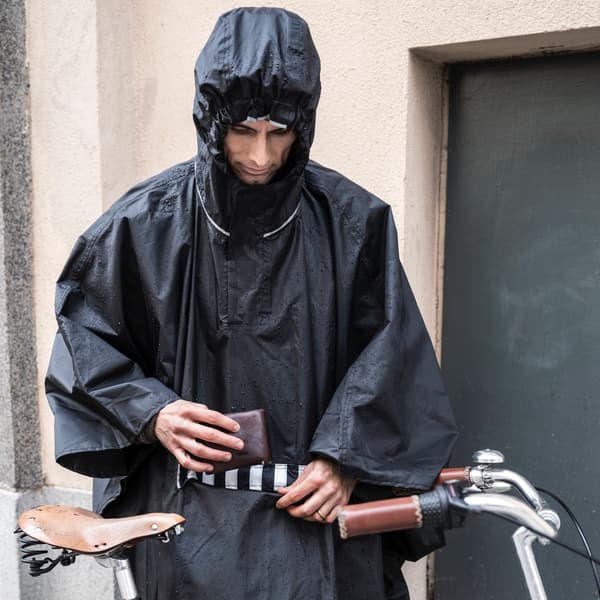 Man in a black hooded rain poncho, adjusting a belt while standing next to a bicycle in an urban outdoor setting.