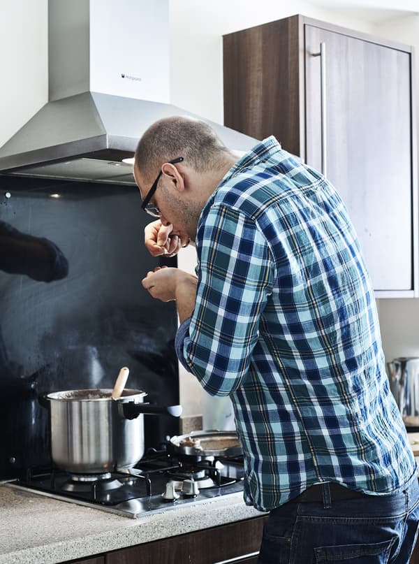 Man in blue shirt is tasting the content of a pot that is standing on the stove.
