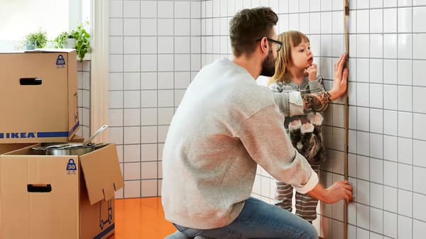 Man measuring wall, daughter in background watching. Moving boxes stacked in corner.