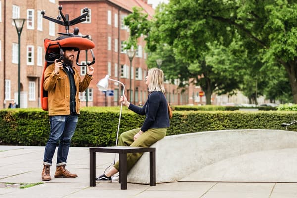 Man med brun jacka står med röd kontorsstol över sitt huvud. Kvinna sitter bredvid på stenbänk med lampa i handen och träbord bredvid.
