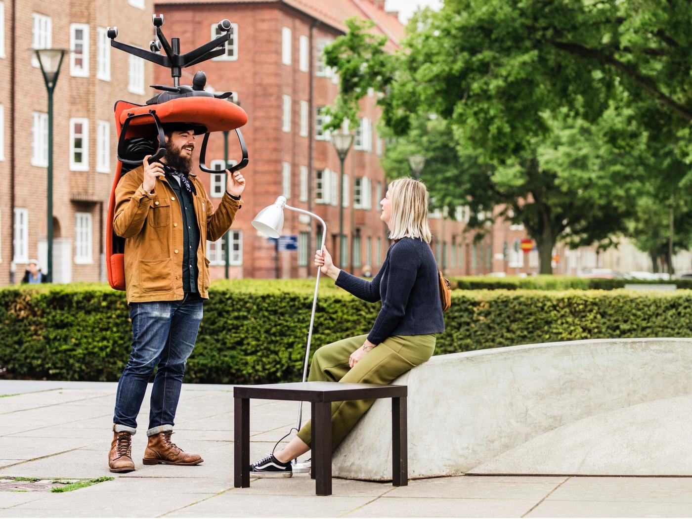 Man med brun jacka står med röd kontorsstol över sitt huvud. Kvinna sitter bredvid på stenbänk med lampa i handen och träbord bredvid.