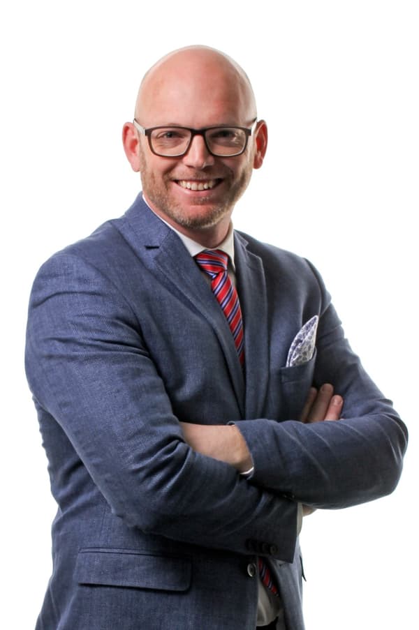 Man med glasögon och blå kostym står med armar i kors och ler