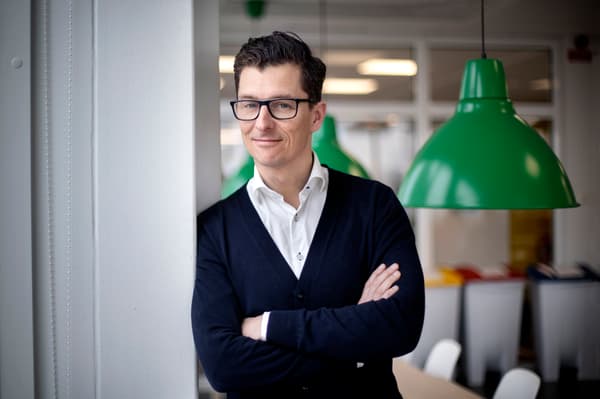 Man med vit skjorta och svart kofta står med armarna i kort och tittar mot kameran