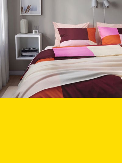 Man sieht ein Schlafzimmer mit einem Doppelbett, rosa karierter Bettwäsche, daneben ein EKET Würfel als Nachttisch und darunter einen gelben Balken.