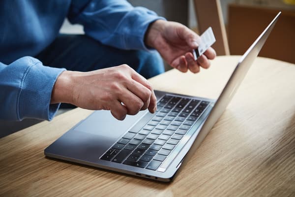 Man sieht einen Laptop und die Hände eine Person. In einer Hand befindet sich eine Kreditkarte.