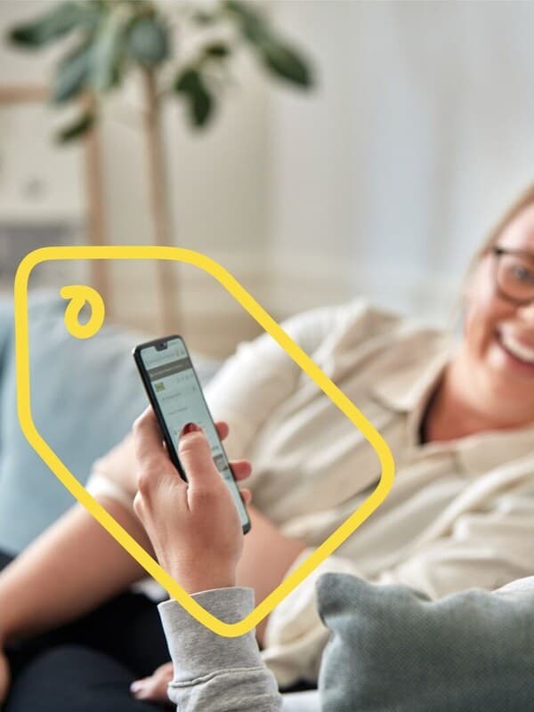 Man sieht im Hintergrund eine lachende Frau am Sofa und davor eine Hand die ein Smartphone hält.