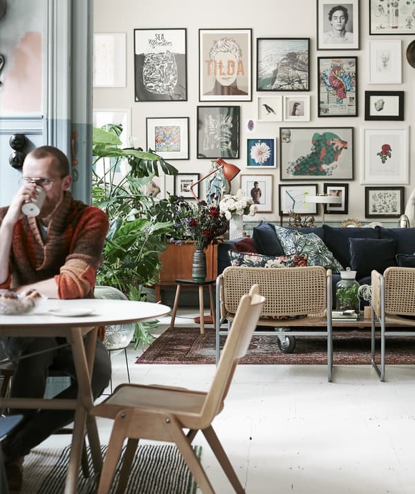 Man sitting at a table drinking from a mug with view of the living room and wall of pictures in the background.