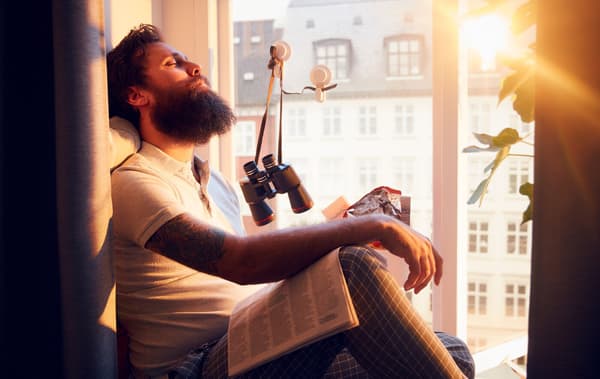 Man sitting on a deep, sunlit window sill, head restfully leaning back. Multistorey facade in the background.