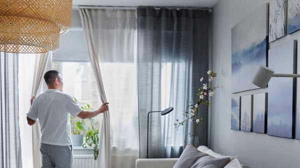 Man som står vid fönster med HILJA gardin i två lager upphängd med gardinsken i taket. 