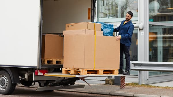 Man unloads large boxes onto pallets from a delivery van in front of a modern building.