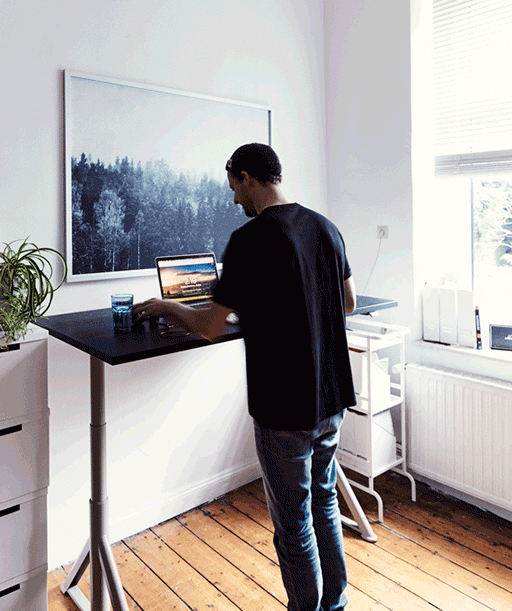 Man using a laptop on an adjustable height desk in a room with white walls and a large picture of a forest.