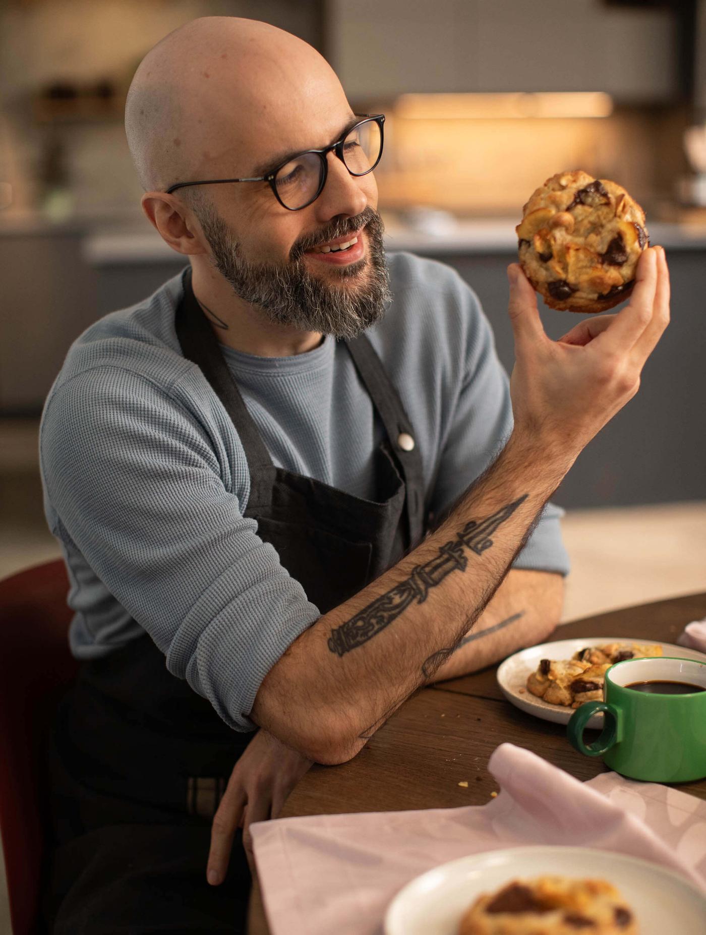 Man wearing glasses and an apron holds up a muffin at a table with coffee and pastries in a kitchen setting.