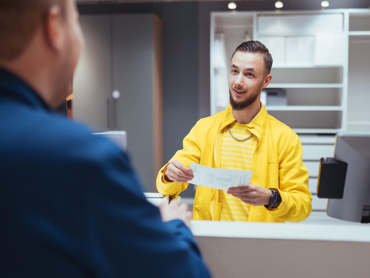 Mand i gul jakke og stribet skjorte står bag en disk og smiler, mens han overrækker et dokument til en anden person. Den anden person er delvist synlig bagfra. Indstillingen, med dens effektive layout og enkle indretning, fremkalder værdien af et IKEA kontor eller receptionsområde.