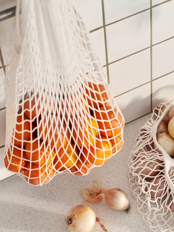 Mandarinnal teli KUNGSFORS hálós táska lóg egy akasztón, mellette a munkalapon egy másik táskában pedig hagymát látunk.
