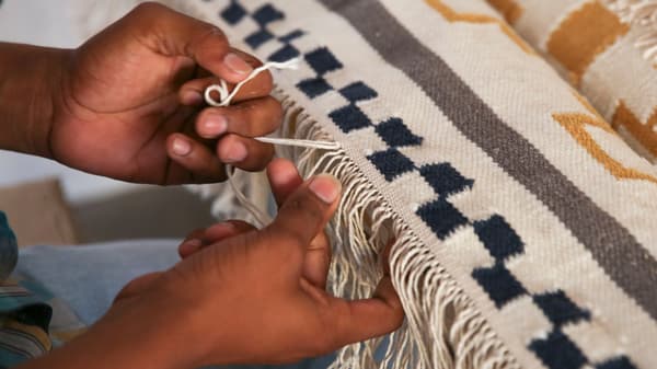 Mani di un artigiano indiano intrecciano la frangia di un tappeto nei toni del bianco, del blu, del grigio e del senape - IKEA.