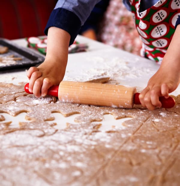 Mani di un bambino che indossa un grembiule e lavora con il mattarello e un po’ di farina l’impasto di pan di zenzero con forme ritagliate.