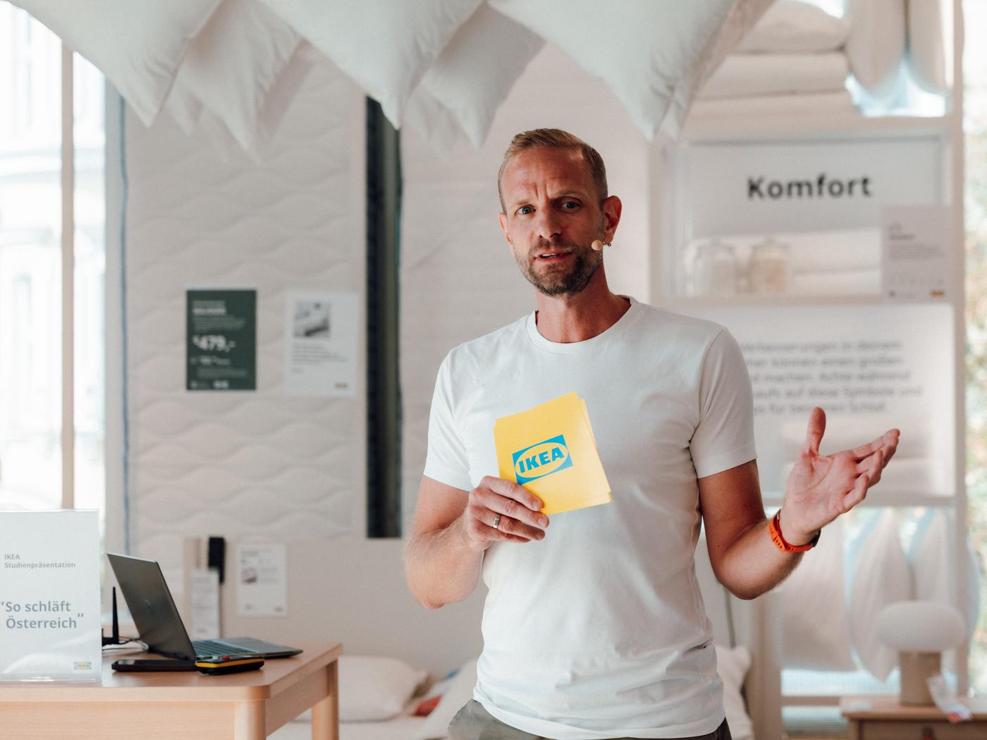 Mann in weißem T-Shirt präsentiert eine IKEA-Karte in einem Schlafbereich mit Matratzen und Kissen