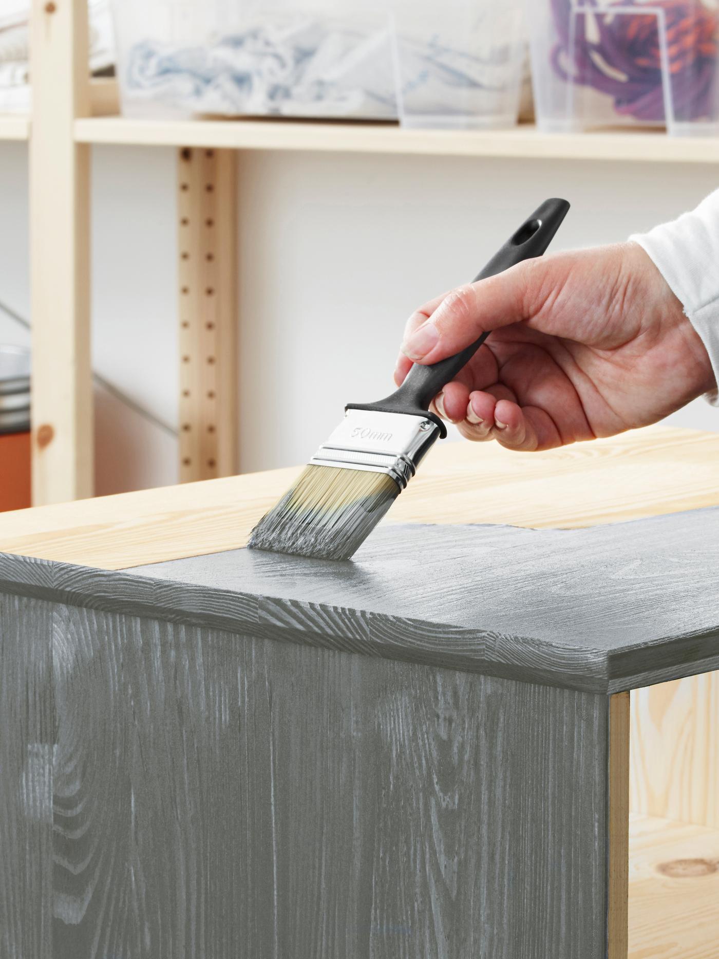 Mano de una persona que sujeta una brocha para aplicar pintura gris a la superficie superior de un mueble de pino sin tratar.