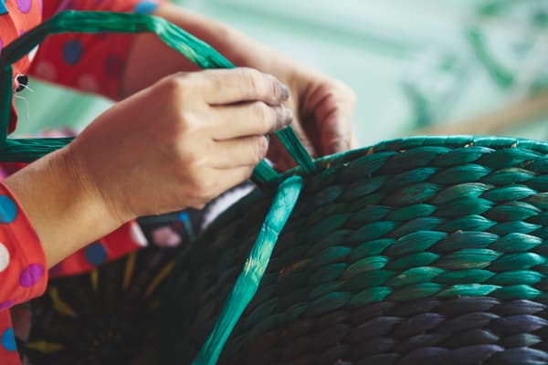Manos armando una cesta de mimbre y fibras naturales de colores negro y verde agua.