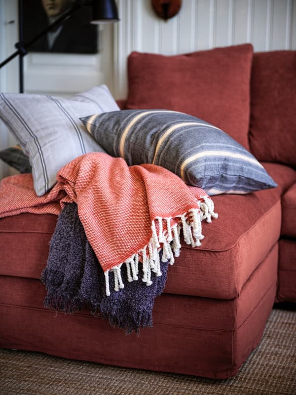 Manta ULLTISTEL roja con bordes blancos sobre otra manta azul. Ambas están sobre un sofá cama IKEA color rojo. Sobre el sofá cama también hay cojines con fundas de textura y lisos.