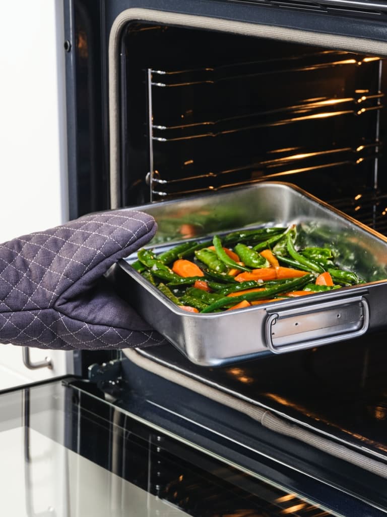 Mão com luva a colocar um tabuleiro para forno KONCIS com cenouras e outros vegetais num forno.