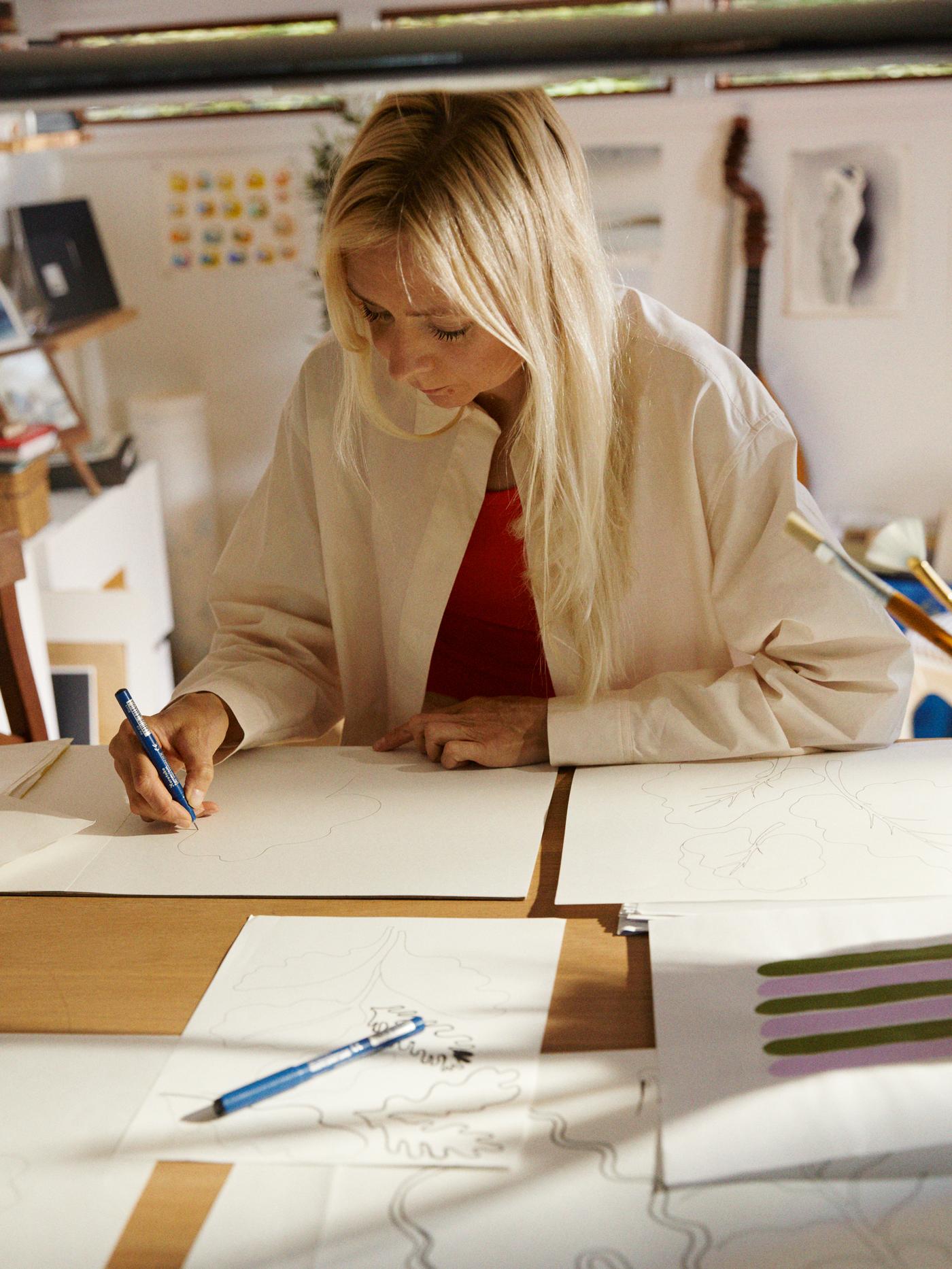 햇살이 내리쬐는 작업실 책상에 놓인 종이에 패턴을 스케치하고 있는 Marimekko 디자이너 Maija Louekari의 모습.
