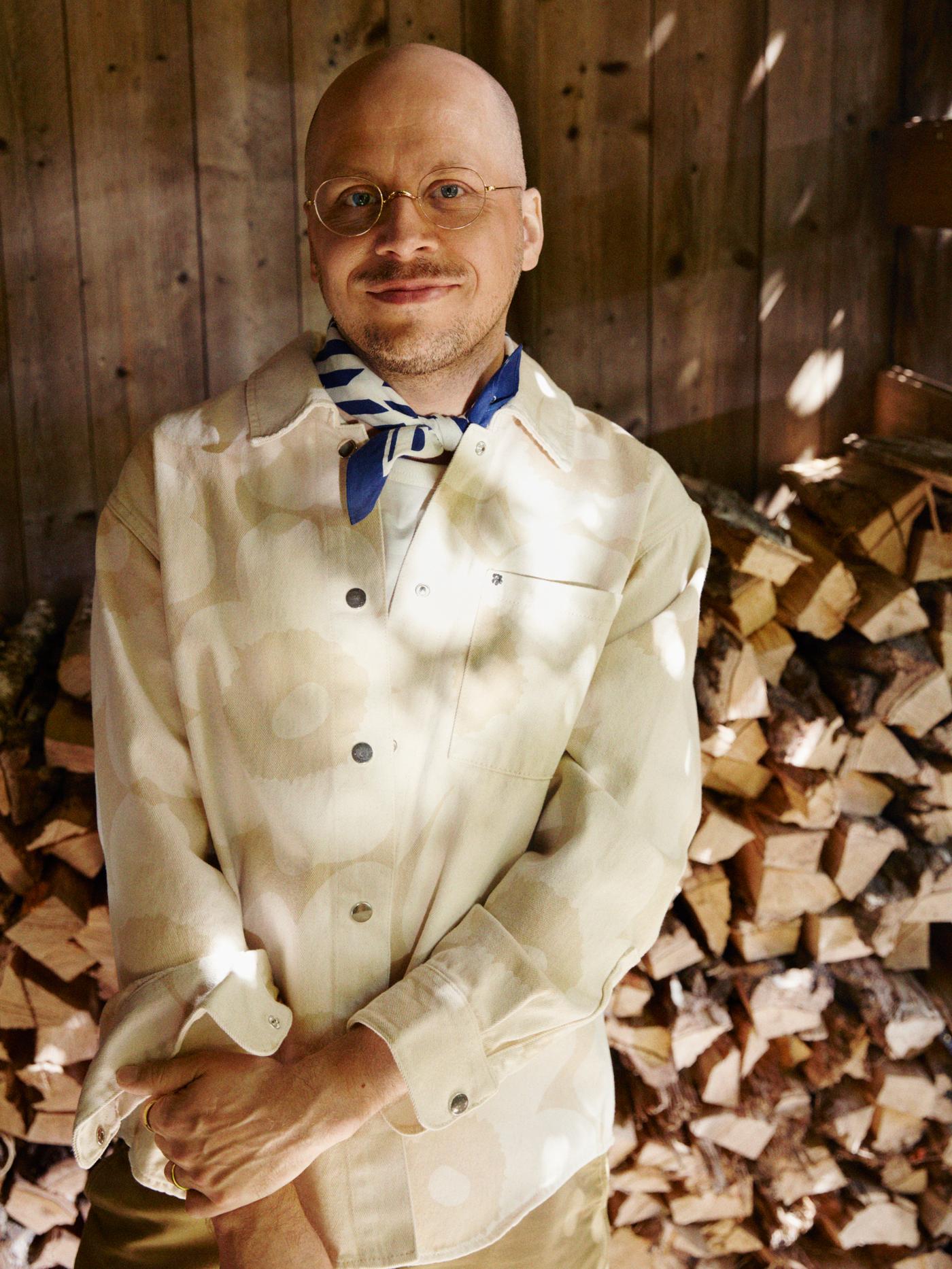 Marimekko의 디자이너 Sami Ruotsalainen이 플로럴 패턴 재킷을 입고 수북이 쌓여 있는 땔감 나무 앞에 서 있는 모습.