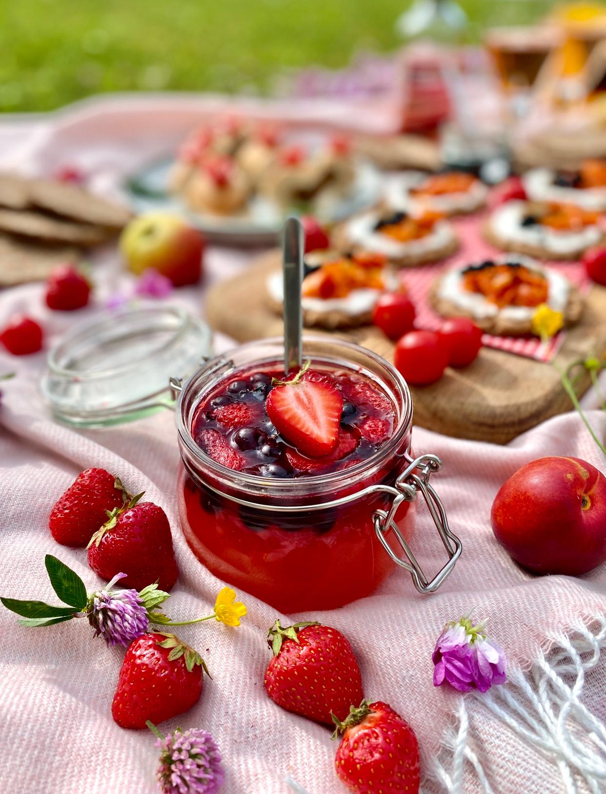 Marmellata di fragole fresche in un barattolo di vetro con un cucchiaio, circondato da fragole intere e da uno sfondo da picnic.