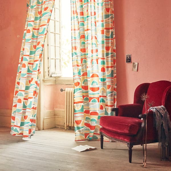 MARTORN curtains with a multi-coloured graphic pattern blowing in the wind of an open window next to an armchair.