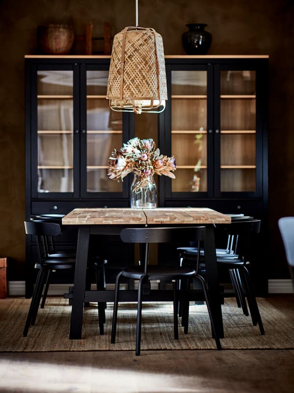 Masă SKOGSTA cu vază cu flori sub o lustră KNIXHULT, înconjurată de scaune de culoare închisă. Corpuri închise la culoare la perete.