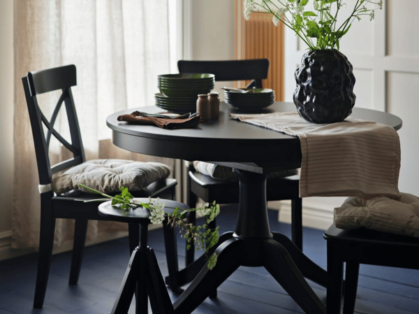 Matbord och stolar i svartbetsat trä står i ett ljust rum med vita tunna gardiner och ett blått trägolv. En svart vas med blommor i står på bordet.