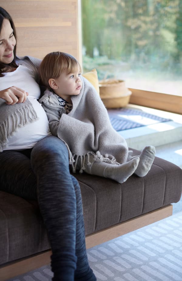 Matka s malým dieťaťom zabaleným v sivej vlnenej priskrývke OMTÄNKSAM sedia spolu na pohovke.