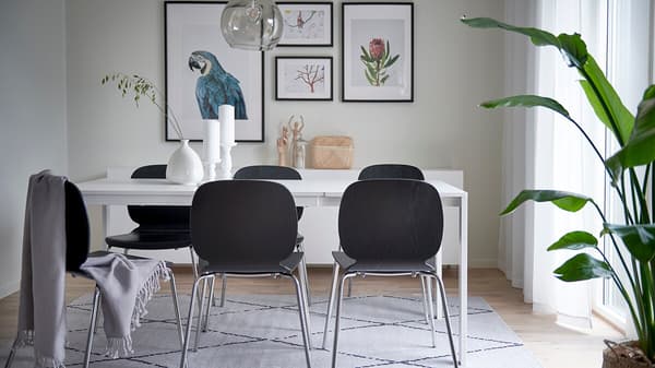 Matplats med vitt matbord och mörkgrå stoppade stolar med metallben placerade på vit matta med diskret, svart mönster. På den ljusgrå väggen bakom bordet finns ett vitt, lågt skåp med fyra tavlor i svarta ramar ovanför. 